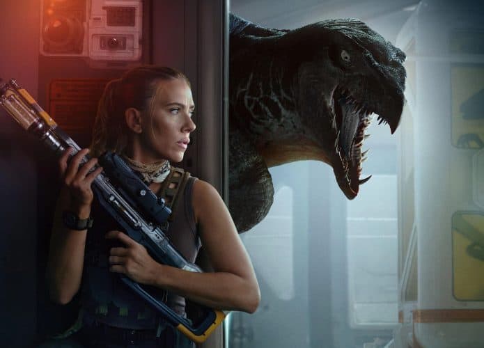 mulher loira com uma arma se escondendo em uma parede com um dinossauro no fundo