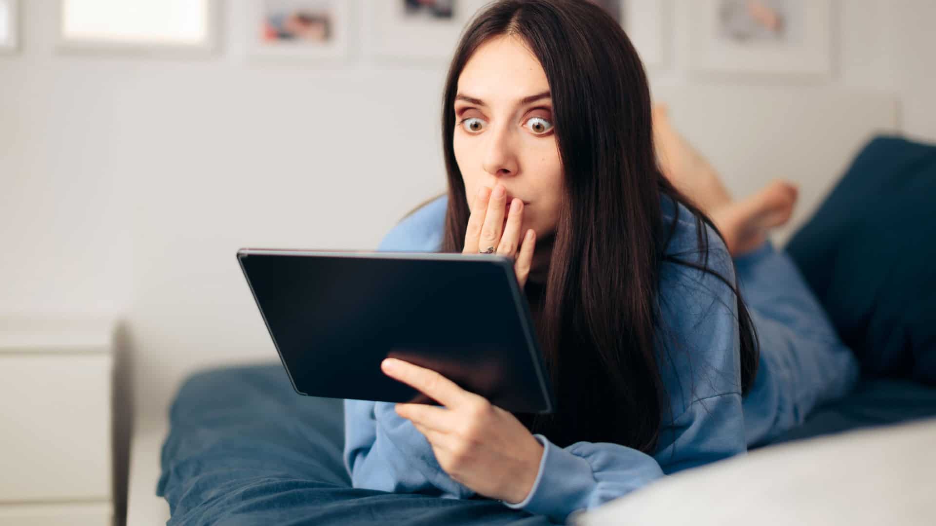 Mulher surpresa deitada em cima da cama e com o tablet na mão
