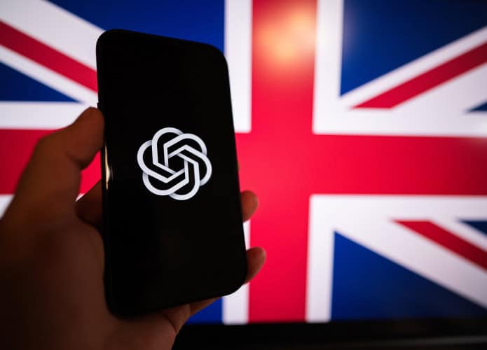 Ao fundo, bandeira do Reino Unido; à frente, uma pessoa segura um smartphone com o logo da OpenAI
