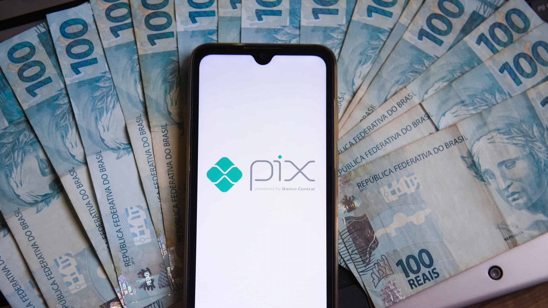 Notas de R$ 100 espalhadas; por cima, um smartphone com o logotipo do Pix