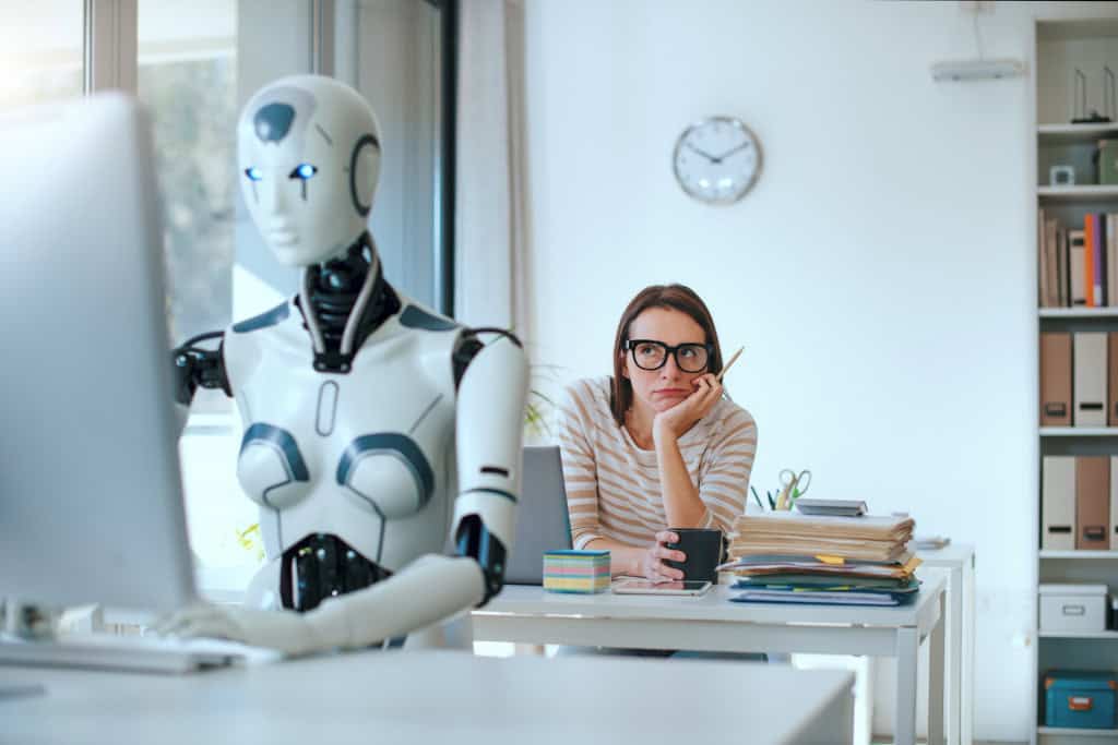 Robô, em primeiro plano, digitando em um teclado de computador; atrás, uma mulher olhando o robô com cara entediada
