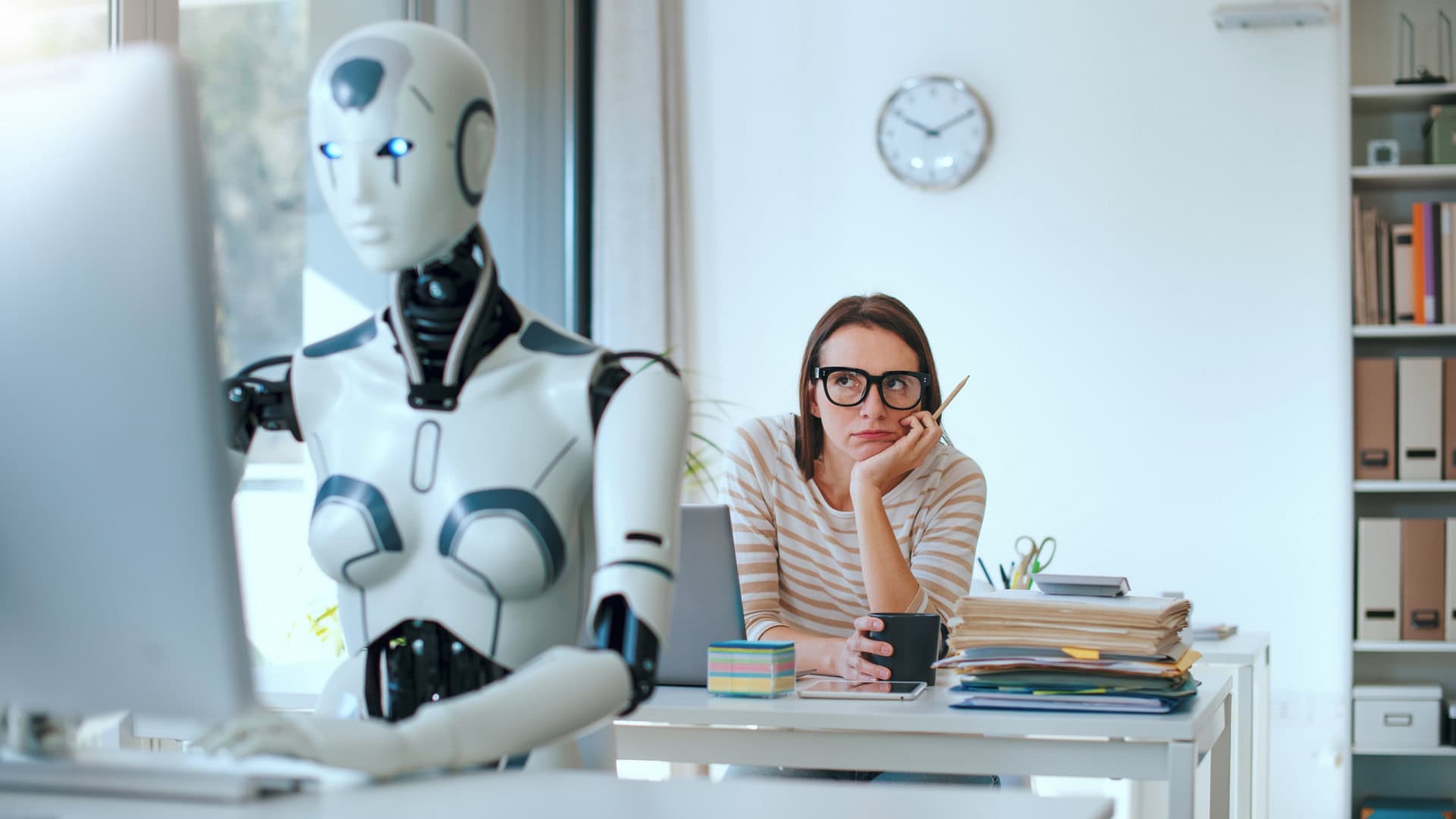 Robô, em primeiro plano, digitando em um teclado de computador; atrás, uma mulher olhando o robô com cara entediada