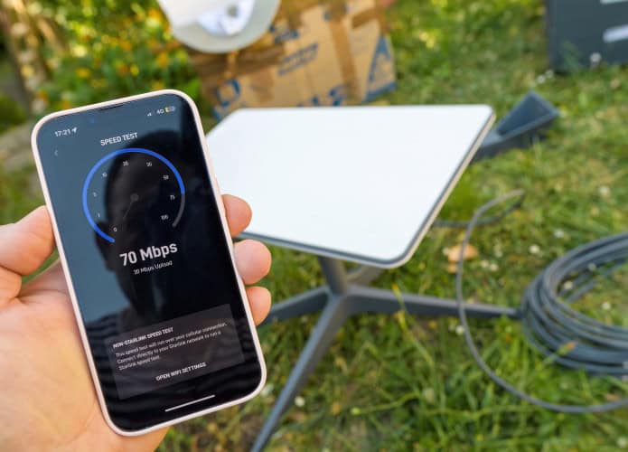 Ao fundo, antena da Starlink em um gramado; acima, pessoa segura um smartphone medindo a velocidade da internet