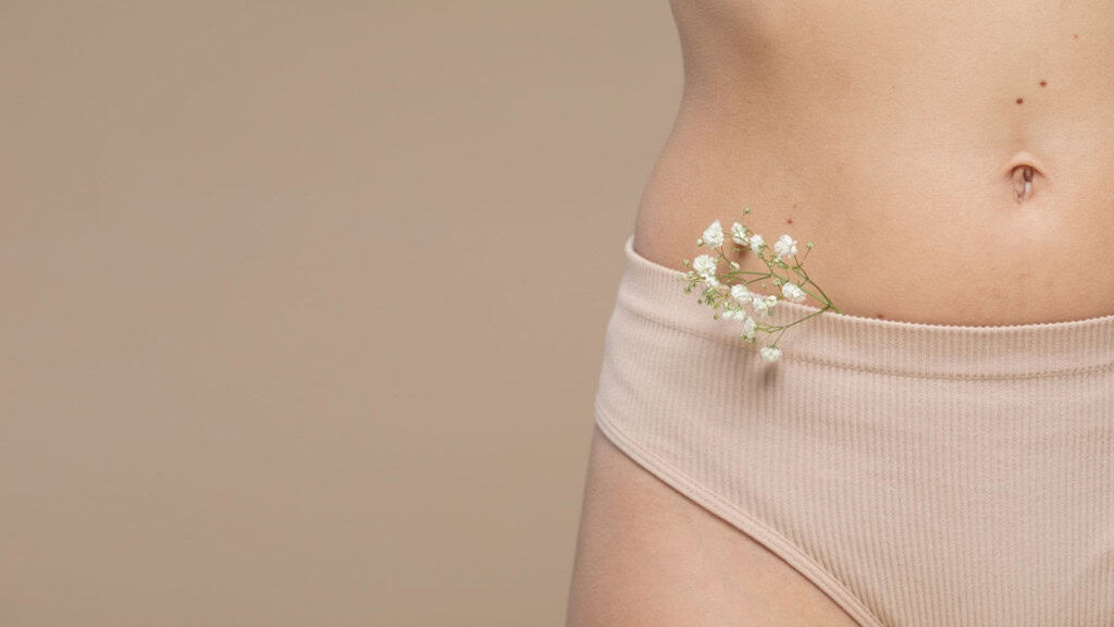 A imagem mostra o abdômen inferior de uma pessoa usando uma calcinha bege. Pequenas flores brancas estão delicadamente posicionadas na borda da peça íntima. O visual mistura suavidade natural com elementos femininos, criando uma composição estética e sutil.