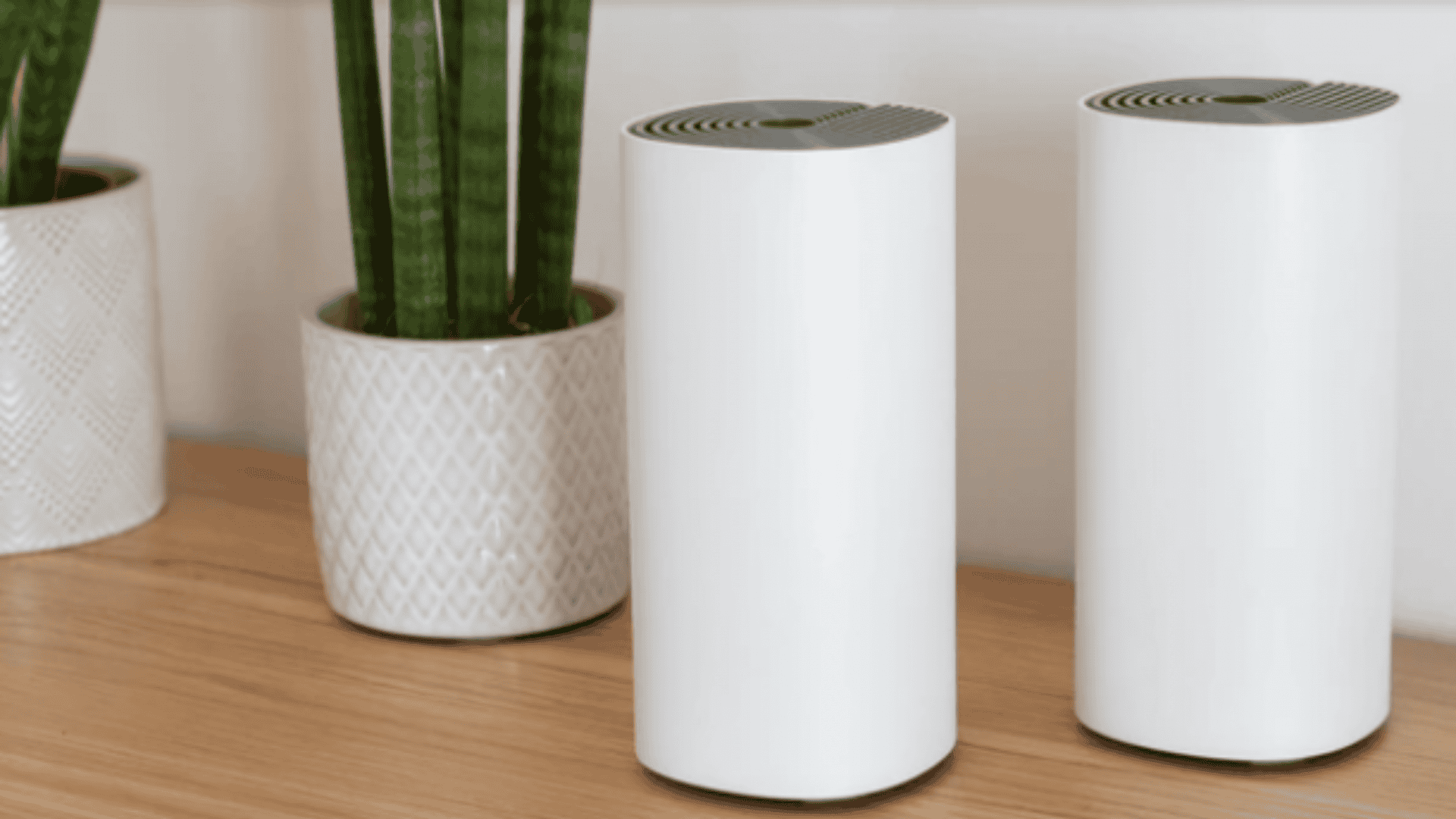 roteadores mesh em cima de uma mesa de madeira, com um vaso de plantas ao lado e uma parede branca atrás