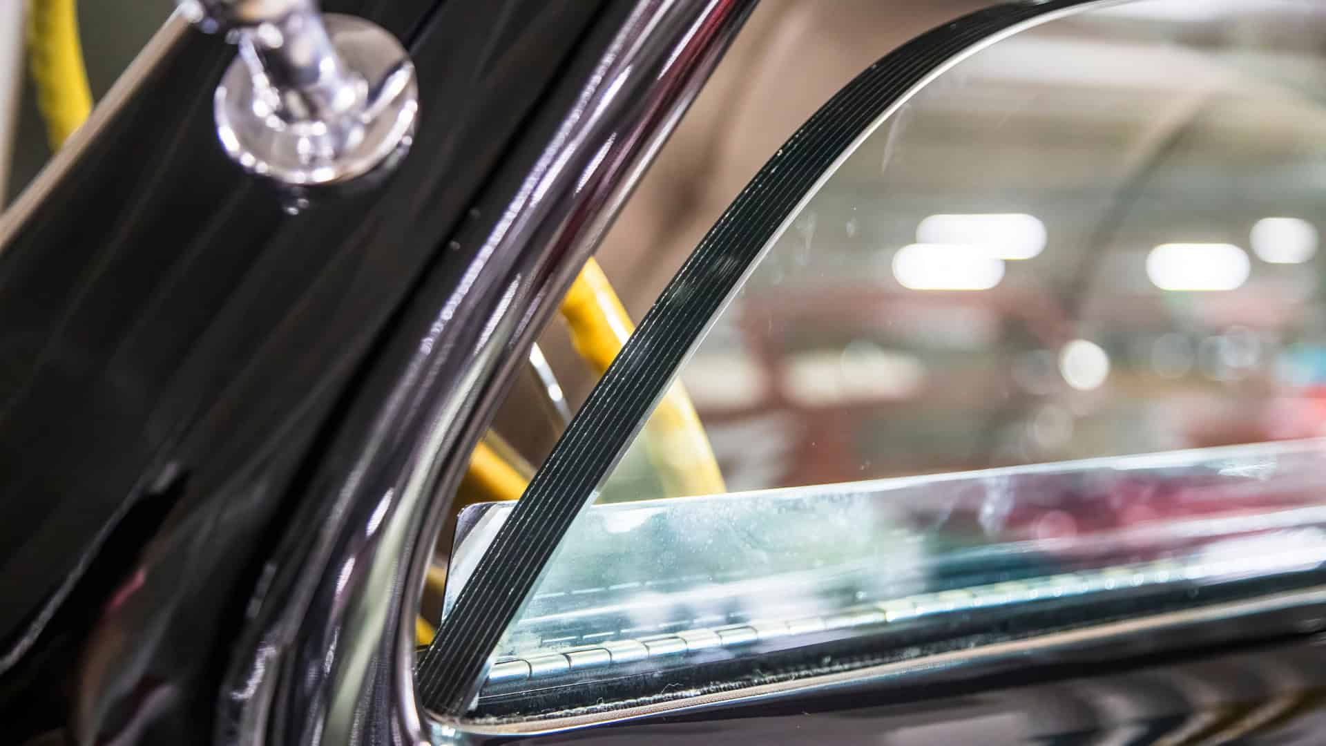 Close-up de uma janela de carro blindado com vidro reforçado espesso, borracha weatherstripping, e acabamento cromado, refletindo a iluminação showroom