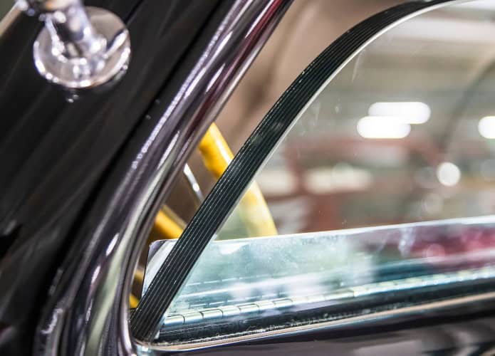 Close-up de uma janela de carro blindado com vidro reforçado espesso, borracha weatherstripping, e acabamento cromado, refletindo a iluminação showroom
