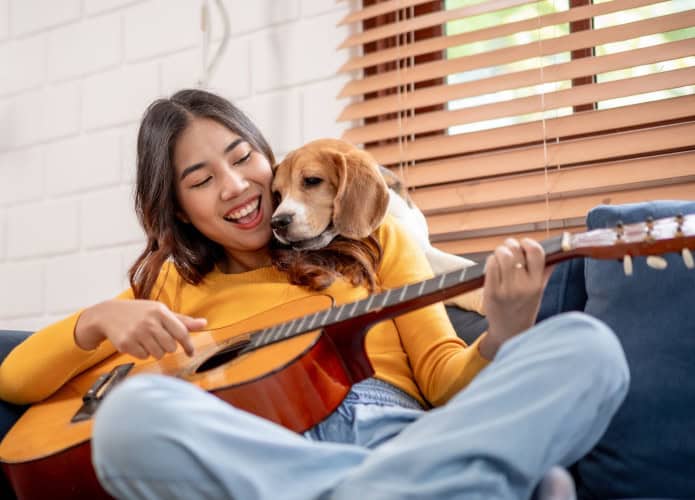 Mulher asiática atraente brinca com seu cachorro beagle na sala de estar de casa. Jovem dona sente-se feliz e relaxada, aproveita para tocar violão e passar momentos de lazer com seu adorável filhote de estimação dentro de casa.