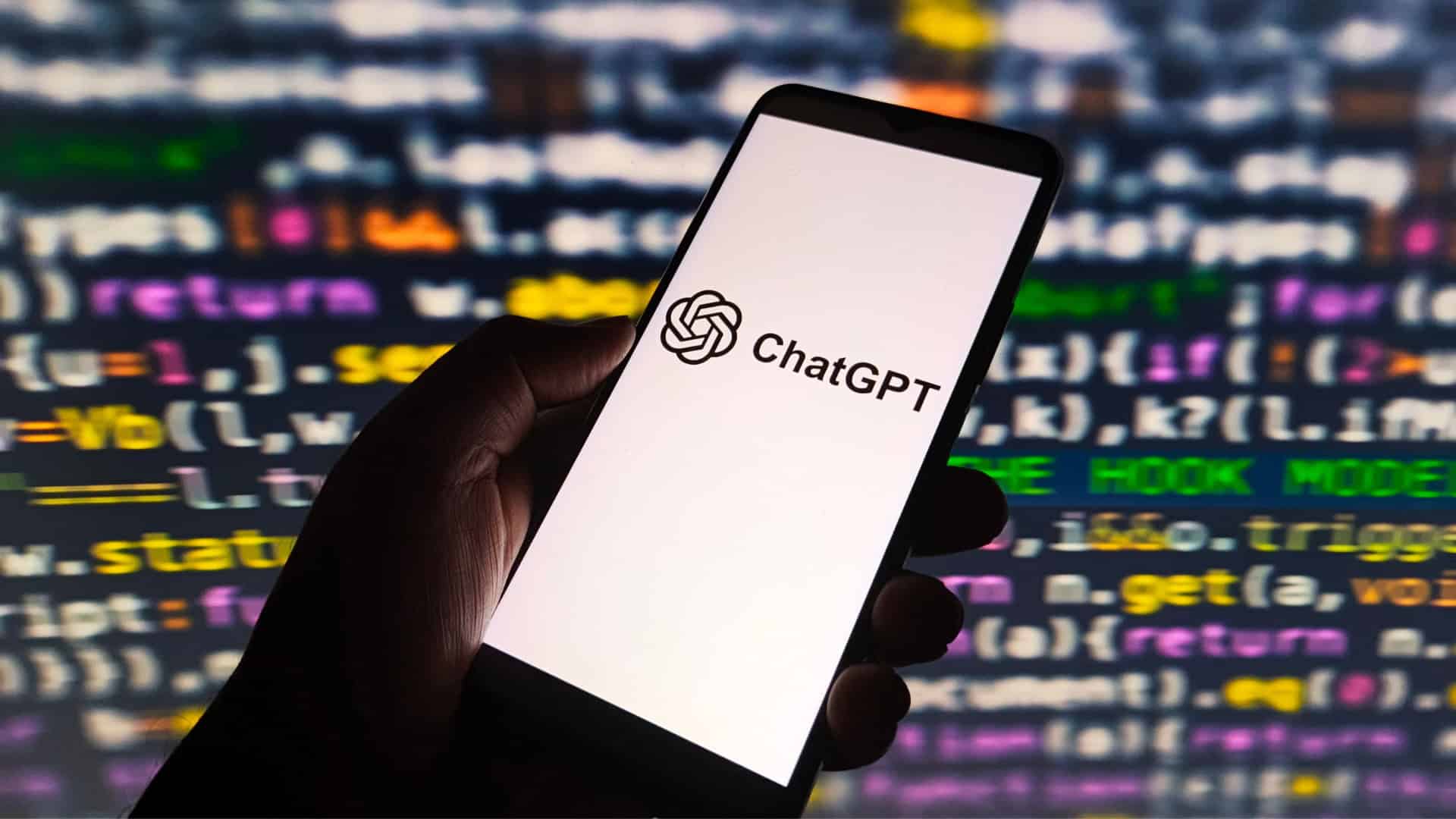Pessoa segurando celular com logotipo do ChatGPT na tela; ao fundo, linhas de código de programação aparecem numa tela grande