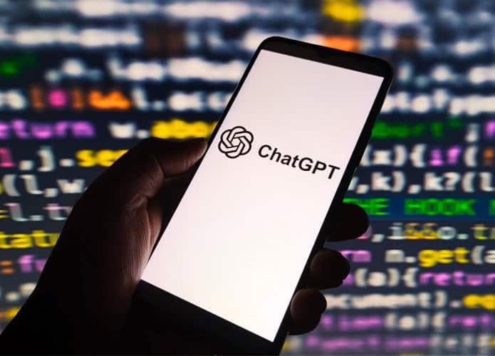 Pessoa segurando celular com logotipo do ChatGPT na tela; ao fundo, linhas de código de programação aparecem numa tela grande