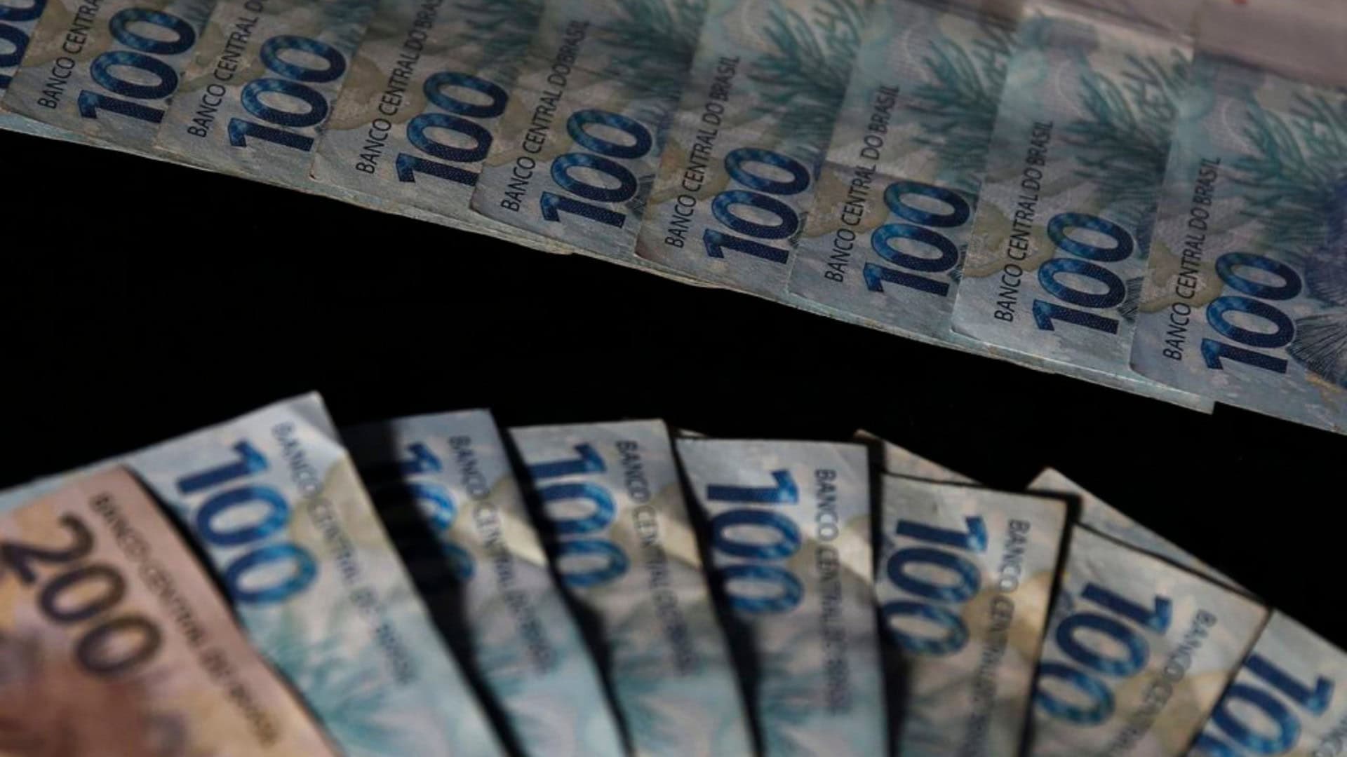Notas de 100 e 200 reais organizadas em formatos de leque e fila numa mesa preta