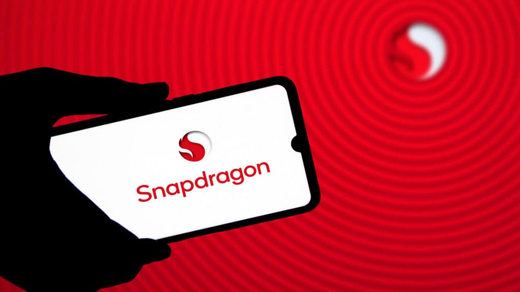 Pessoa segurando celular com logomarca do Snapdragon na tela; ao fundo, uma tela mostrando o logotipo da Qualcomm