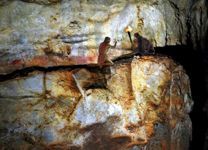 Ilustração de exploradores pré-históricos desenhando em um local perigoso, acima de um precipício de rocha, na Caverna Etxeberri.