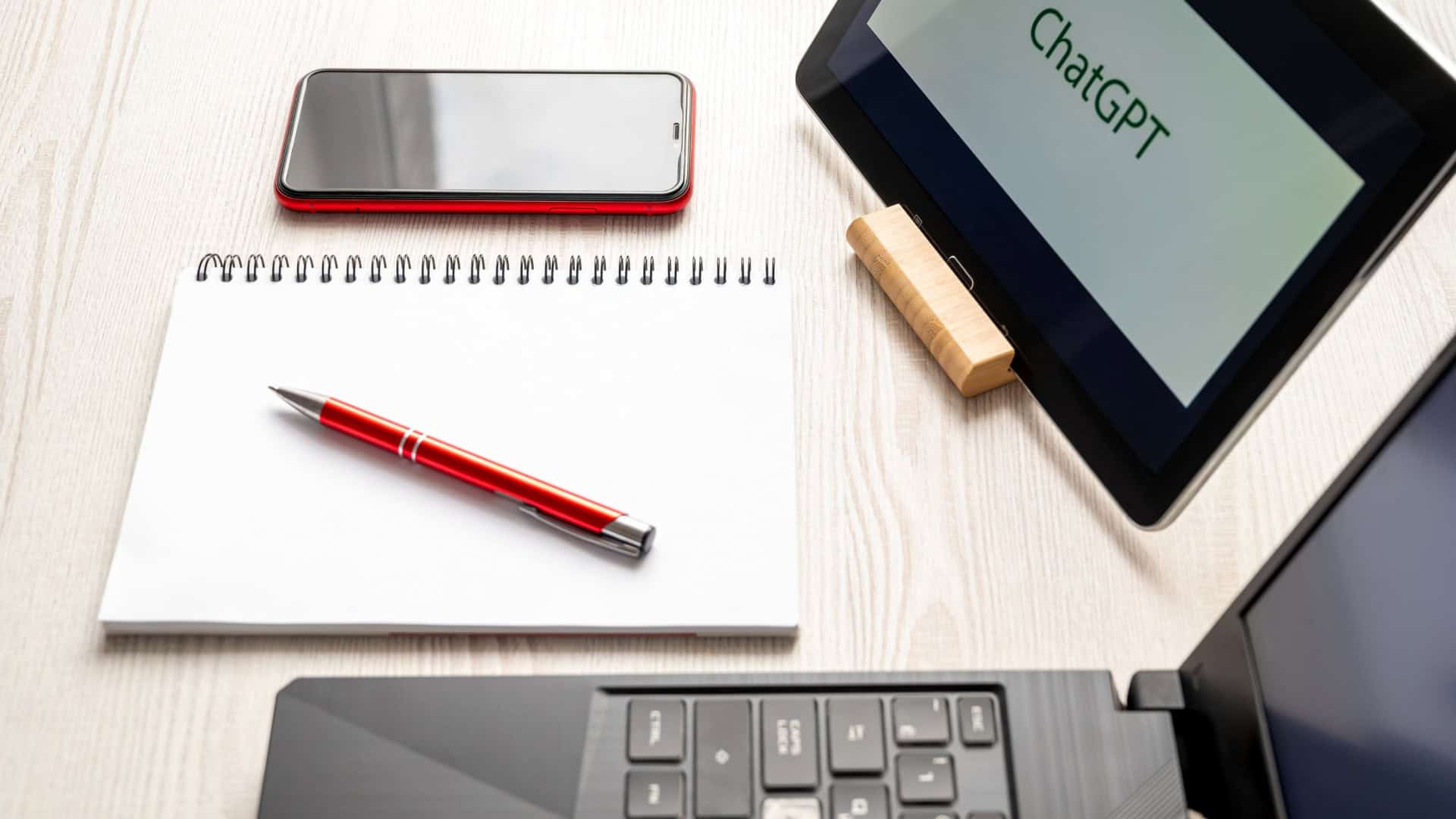 Uma configuração de espaço de trabalho com um laptop, tablet exibindo ChatGPT, smartphone, notebook e caneta em uma mesa de madeira leve.