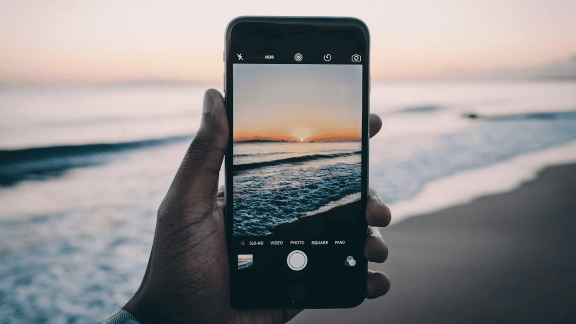 Mão segurando celular com a câmera aberta em frente ao mar