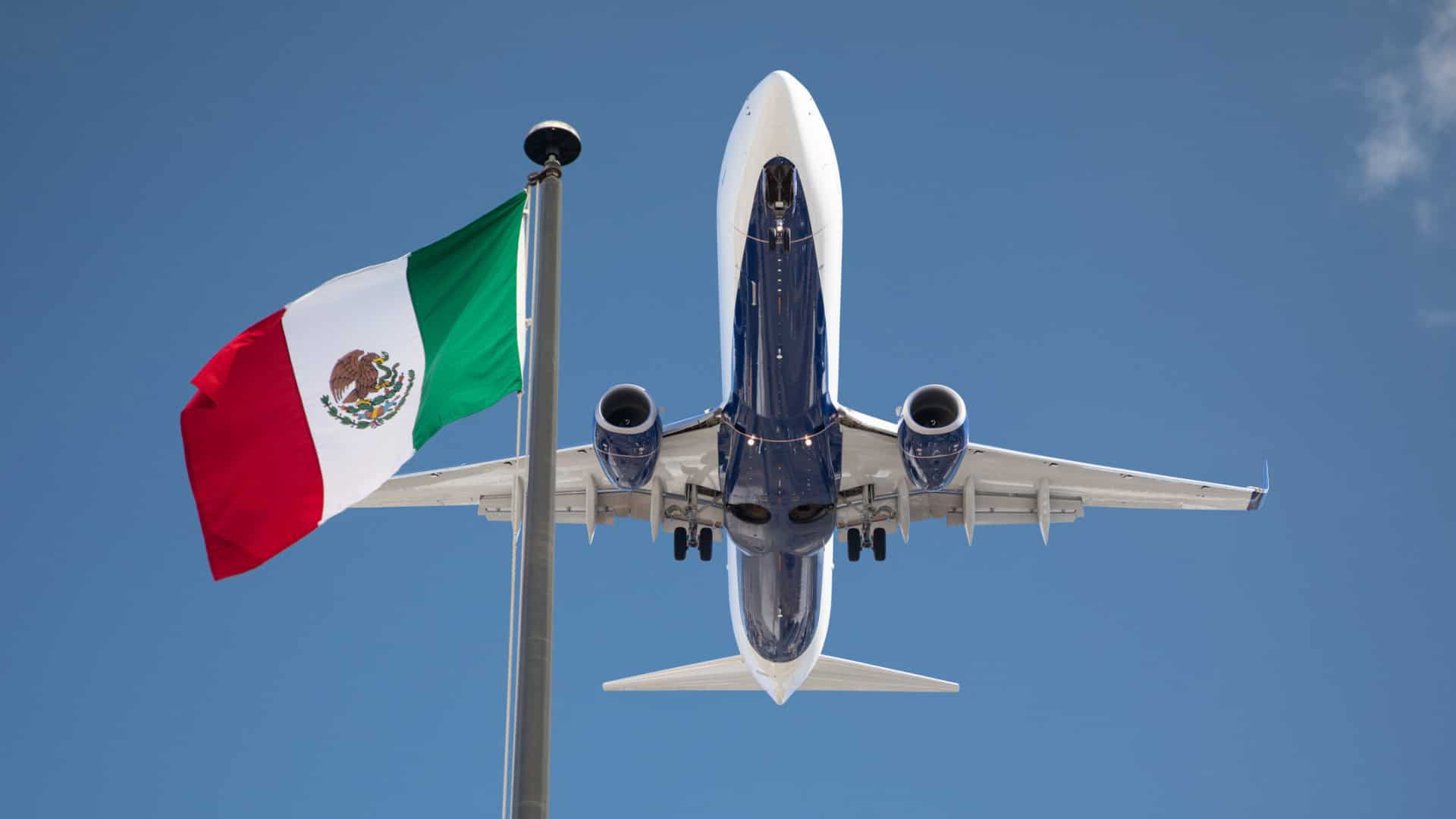 Avião visto de baixo decolando com a bandeira do México em contraste