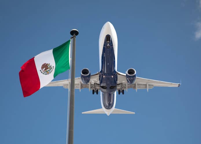 Avião visto de baixo decolando com a bandeira do México em contraste