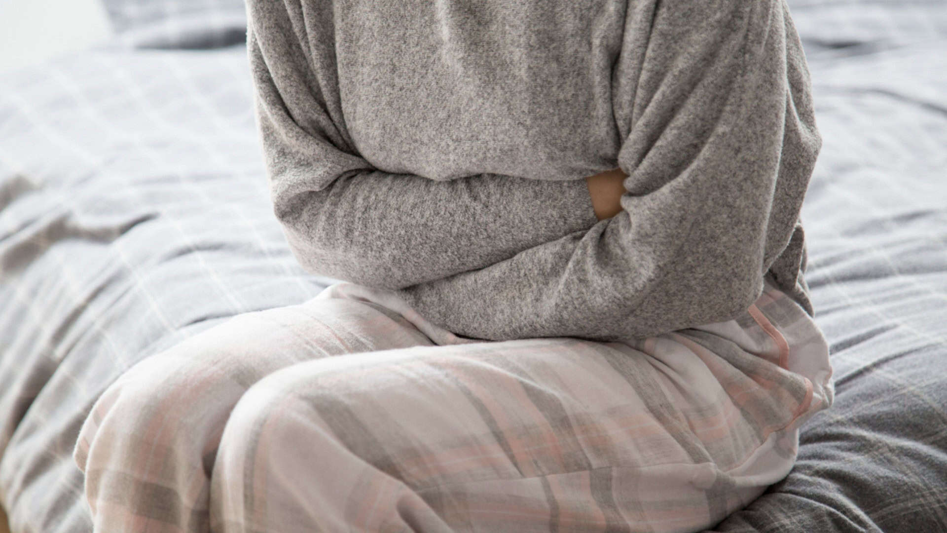 mulher de pijama sentada na cama enquanto aperta a barriga como se sentisse dor
