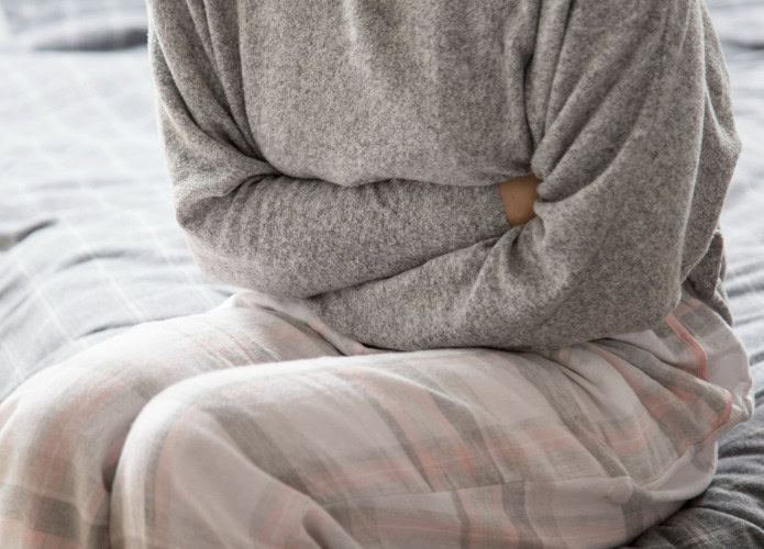 mulher de pijama sentada na cama enquanto aperta a barriga como se sentisse dor