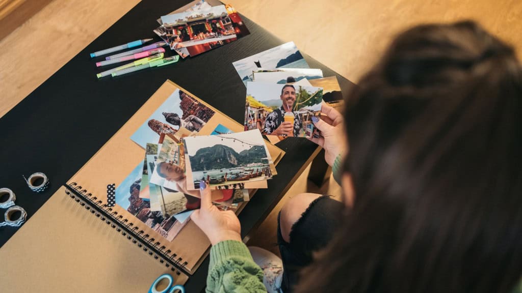 Imagem vista de cima de uma mulher de meia-idade não identificável selecionando fotos para colar com fita washi em seu álbum de viagem artesanal feito de papel kraft.