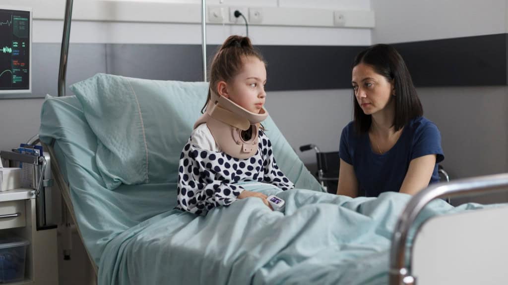 Criança internada em hospital com pescoço imobilizado e mãe ao seu lado.