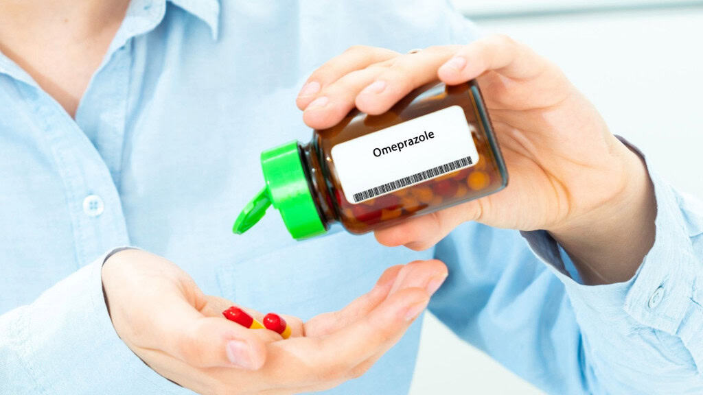 Pessoa com camisa azul segura um frasco marrom com tampa verde escrito “Omeprazol” e despeja cápsulas vermelhas e amarelas na outra mão, representando o uso do medicamento para tratar problemas de acidez no estômago.