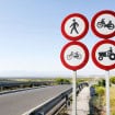 Quatro placas redondas com borda vermelha indicam proibição de pedestres, motos, bicicletas e tratores numa estrada rural cercada por vegetação. Ao fundo, há montanhas sob céu limpo, com guarda-corpos metálicos ao longo da via.