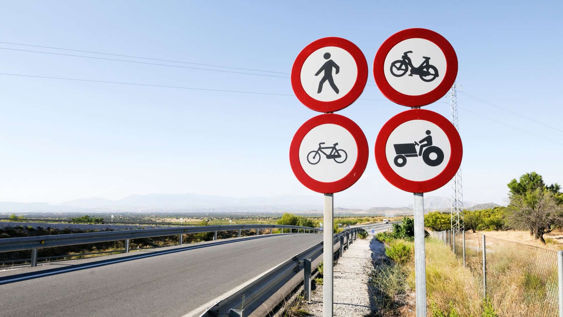 Quatro placas redondas com borda vermelha indicam proibição de pedestres, motos, bicicletas e tratores numa estrada rural cercada por vegetação. Ao fundo, há montanhas sob céu limpo, com guarda-corpos metálicos ao longo da via.