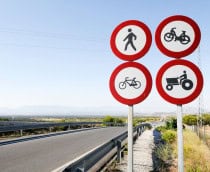 Quatro placas redondas com borda vermelha indicam proibição de pedestres, motos, bicicletas e tratores numa estrada rural cercada por vegetação. Ao fundo, há montanhas sob céu limpo, com guarda-corpos metálicos ao longo da via.