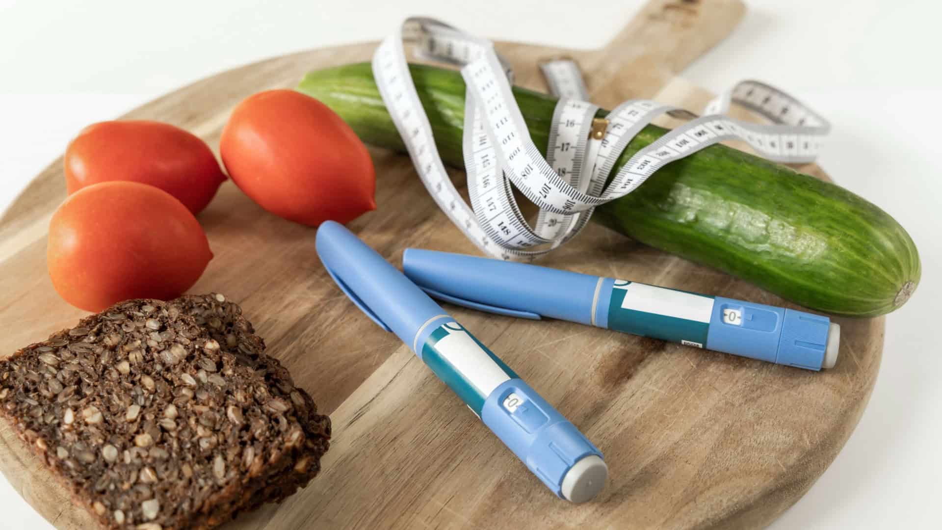 Sobre uma tábua de madeira estão três tomates vermelhos, uma fatia de pão integral, um pepino com fita métrica enrolada, e duas canetas de insulina azuis. A cena mistura alimentos saudáveis com itens médicos, sugerindo controle da diabetes por meio da alimentação e medicação.