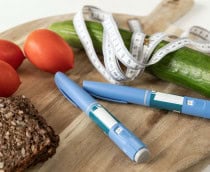 Sobre uma tábua de madeira estão três tomates vermelhos, uma fatia de pão integral, um pepino com fita métrica enrolada, e duas canetas de insulina azuis. A cena mistura alimentos saudáveis com itens médicos, sugerindo controle da diabetes por meio da alimentação e medicação.