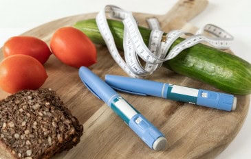 Sobre uma tábua de madeira estão três tomates vermelhos, uma fatia de pão integral, um pepino com fita métrica enrolada, e duas canetas de insulina azuis. A cena mistura alimentos saudáveis com itens médicos, sugerindo controle da diabetes por meio da alimentação e medicação.