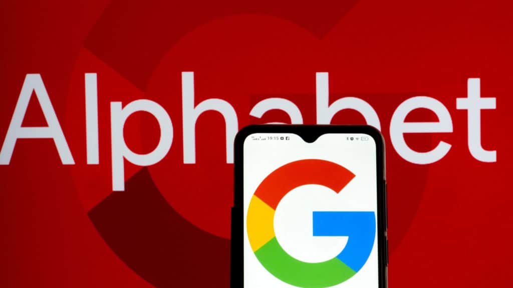 Celular com logotipo do Google na tela colocado na frente de uma tela exibindo letreiro escrito Alphabet