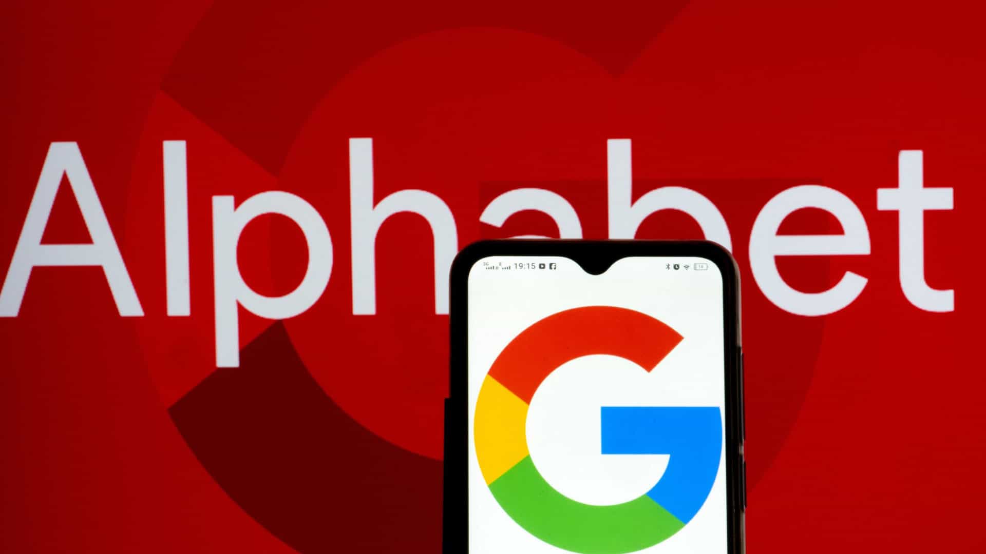 Celular com logotipo do Google na tela colocado na frente de uma tela exibindo letreiro escrito Alphabet
