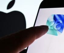 Dedo de pessoa quase tocando ícone do iOS 26 em tela de celular colocado na frente de tela maior exibindo o logotipo da Apple