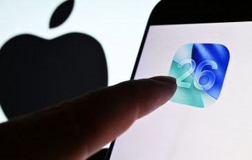 Dedo de pessoa quase tocando ícone do iOS 26 em tela de celular colocado na frente de tela maior exibindo o logotipo da Apple