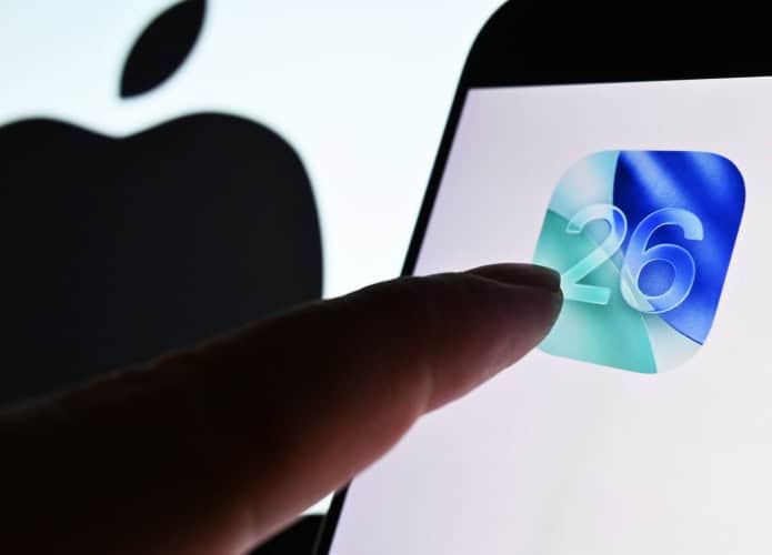 Dedo de pessoa quase tocando ícone do iOS 26 em tela de celular colocado na frente de tela maior exibindo o logotipo da Apple