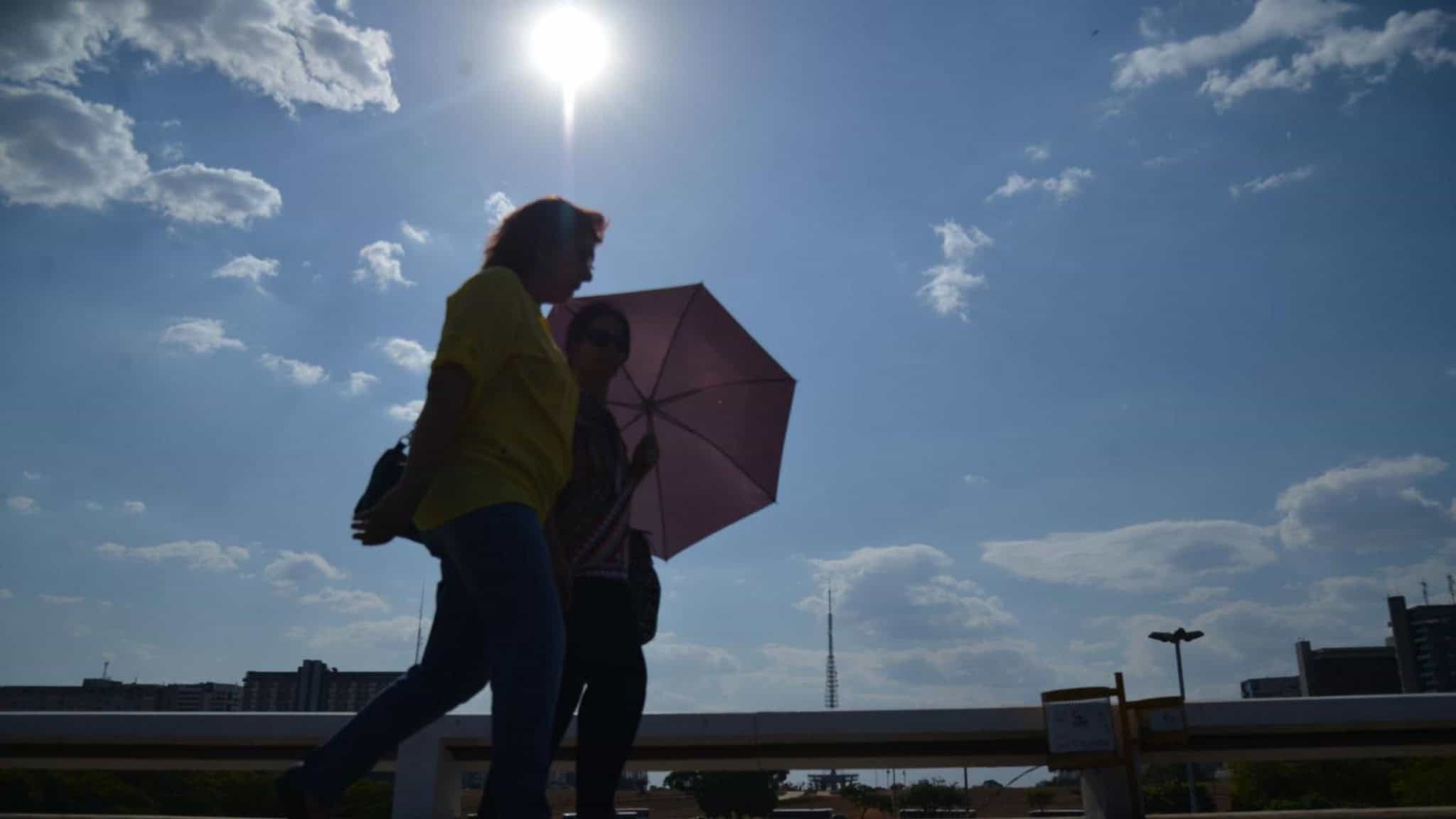 Duas mulheres andando na rua. Uma delas está com uma sombrinha rosa sobre a cabeça. O dia está ensolarado e parece quente.
