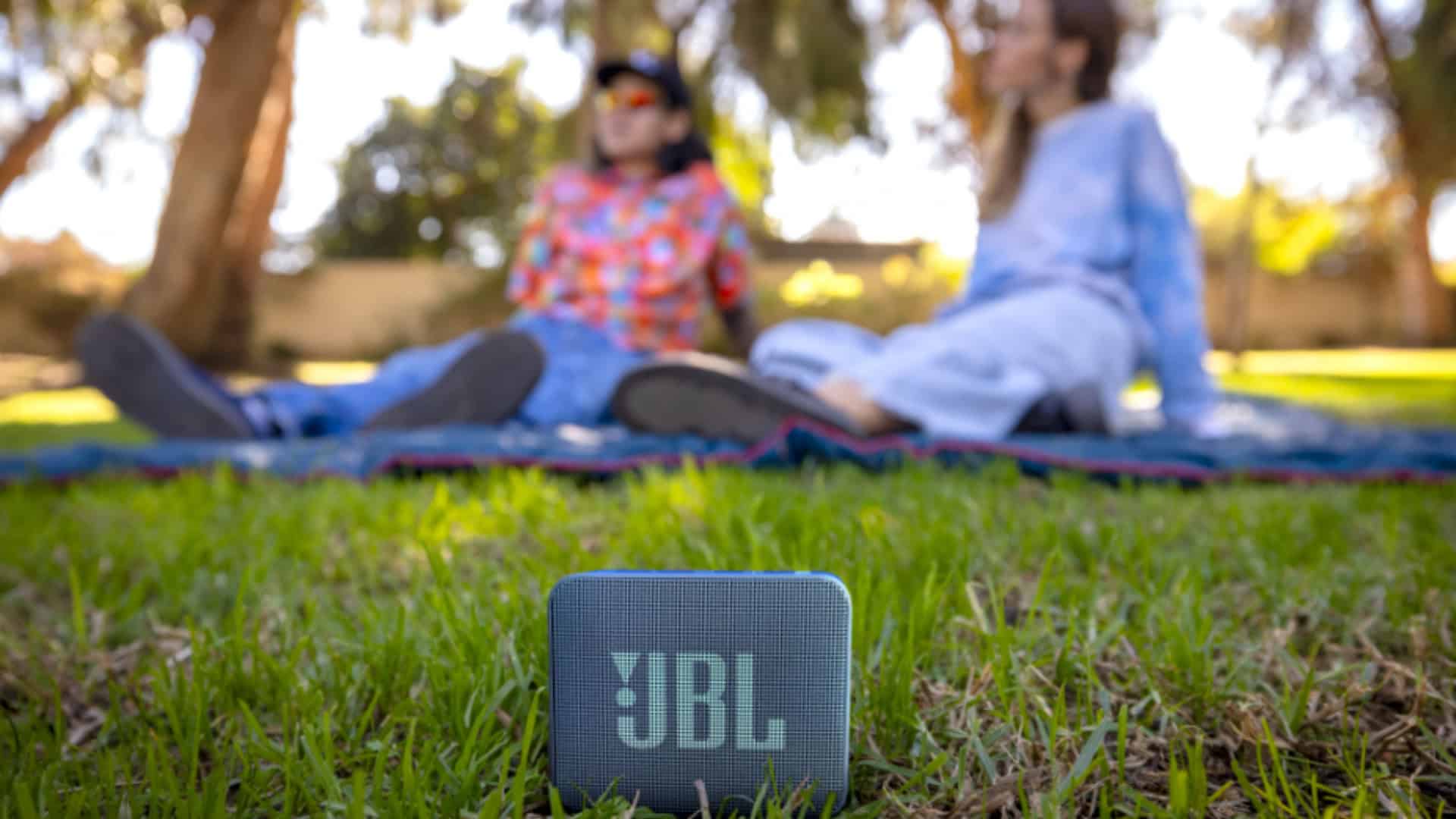 Nova caixinha JBL Go Essential 2 na grama na frente de duas moças sentadas em cima de toalha num parque
