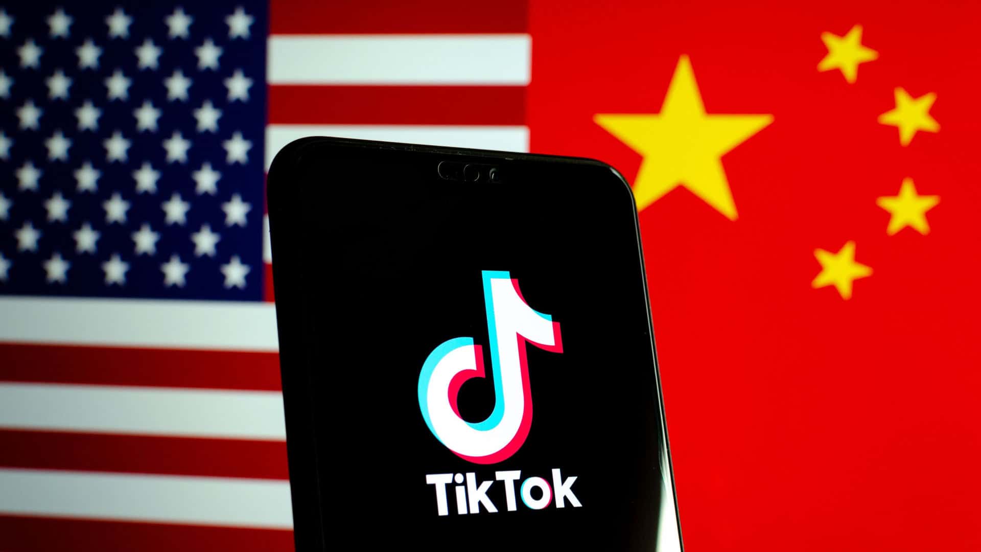 Celular com logotipo do TikTok na tela colocado na frente de uma tela com uma imagem que metade é a bandeira dos Estados Unidos e metade é da China