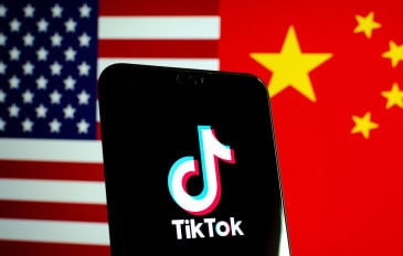 Celular com logotipo do TikTok na tela colocado na frente de uma tela com uma imagem que metade é a bandeira dos Estados Unidos e metade é da China
