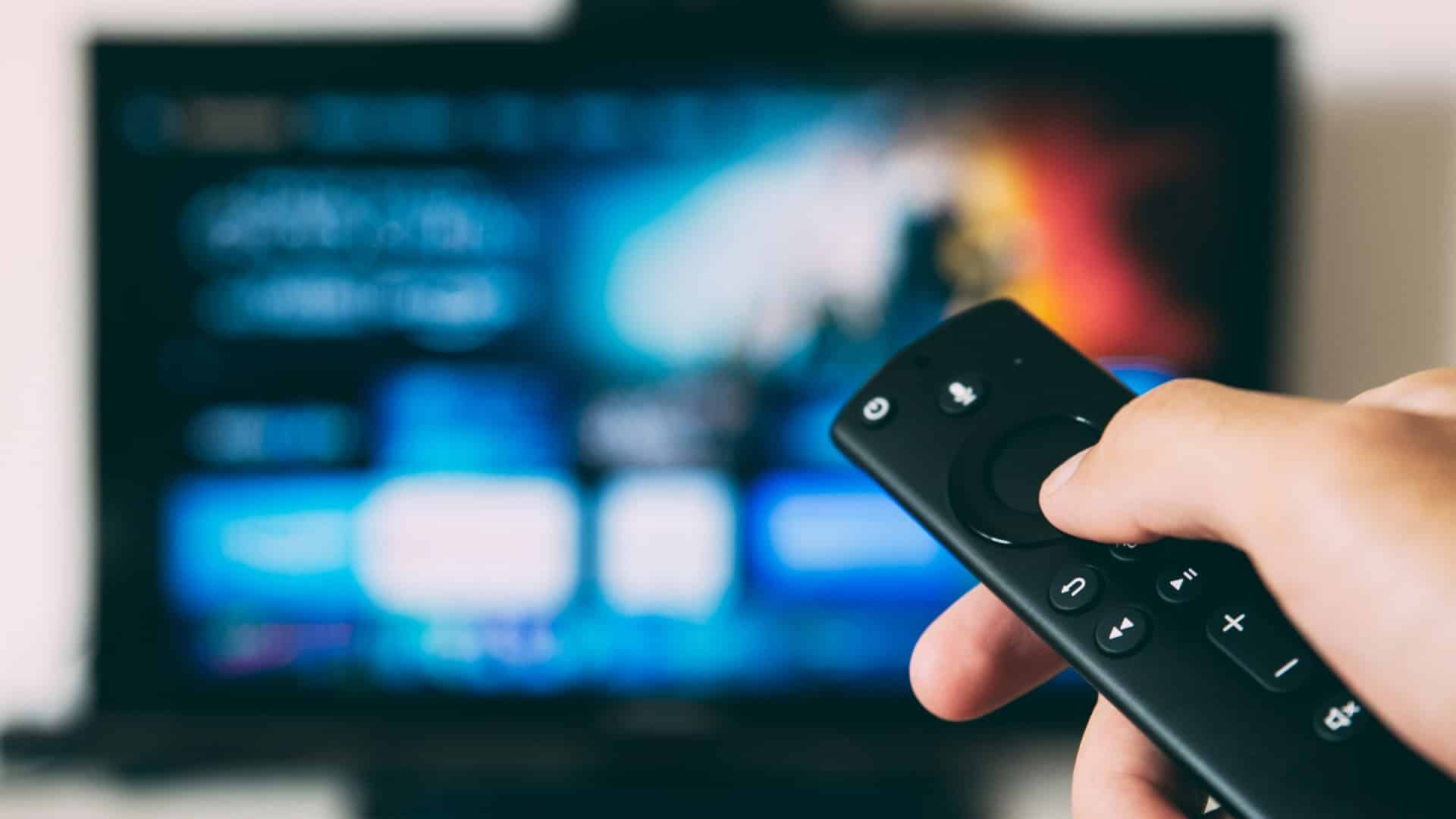 Mão segurando controle com uma televisão ligada ao fundo