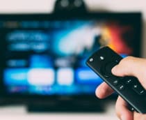 Mão segurando controle com uma televisão ligada ao fundo