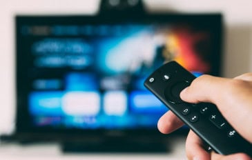 Mão segurando controle com uma televisão ligada ao fundo