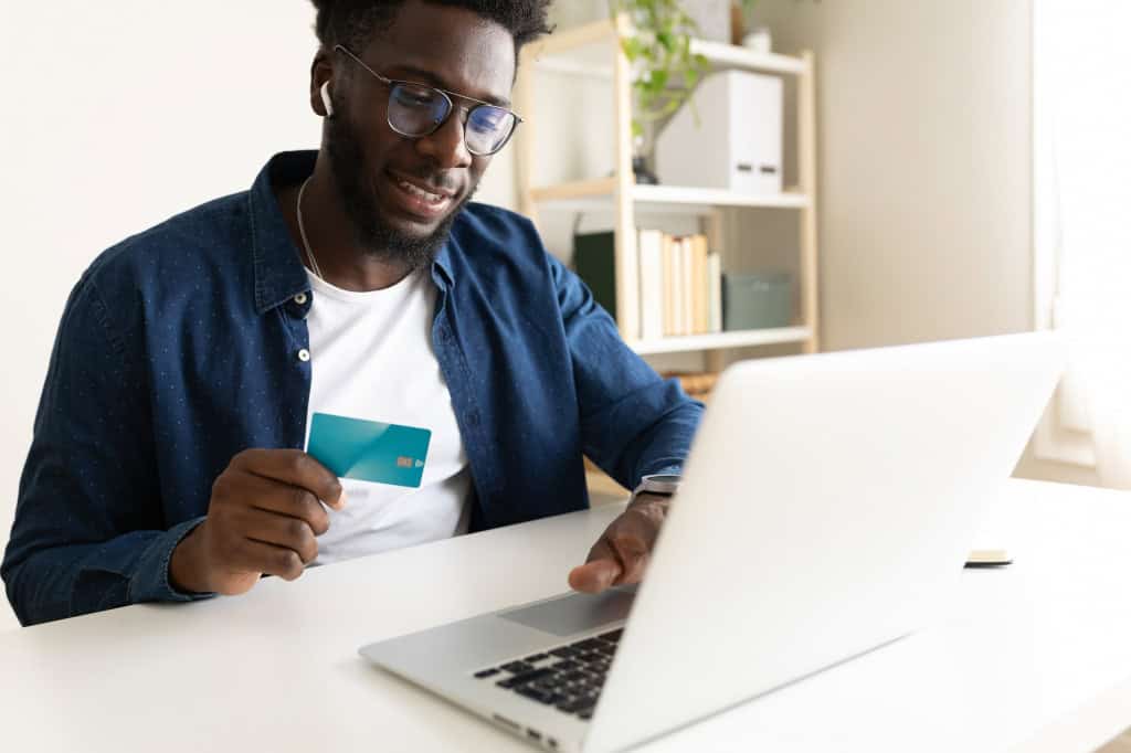 Homem segurando um cartão de crédito com a mão direita e teclando em um notebook com a esquerda