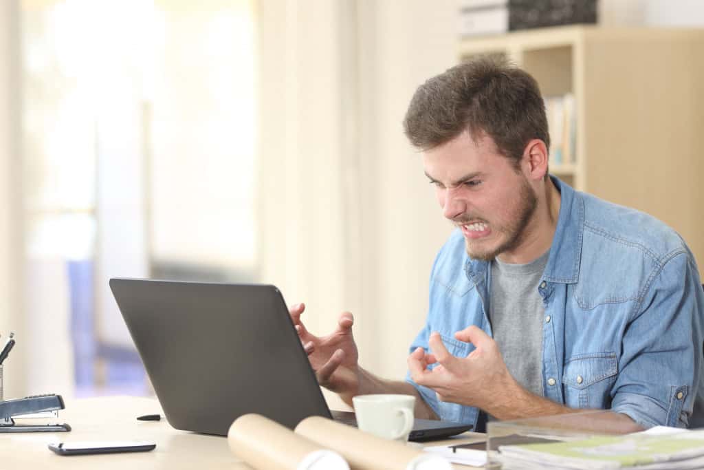 Imagem mostra  homem irritado e furioso com um laptop em um pequeno escritório ou casa