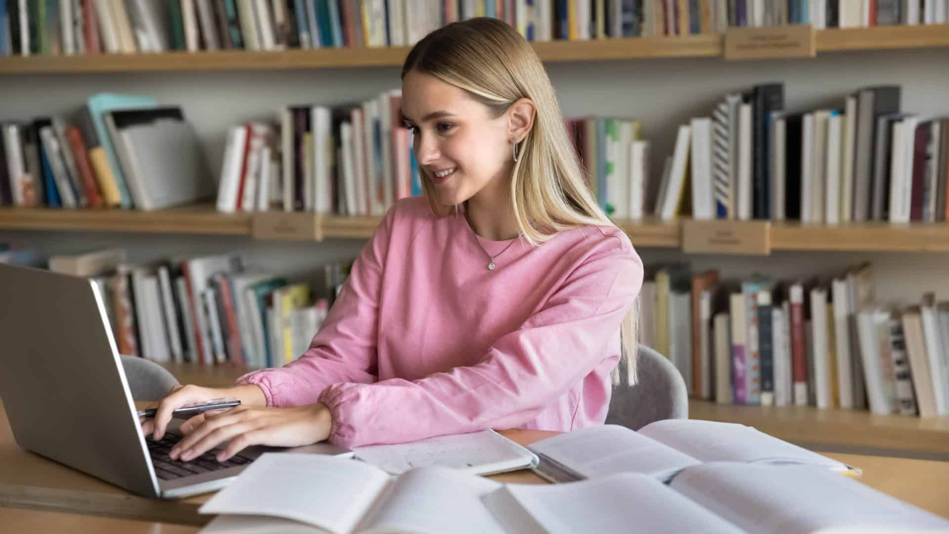 Menina estudando com livros abertos espalhados em uma mesa enquanto digita no notebook