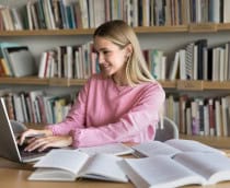 Menina estudando com livros abertos espalhados em uma mesa enquanto digita no notebook
