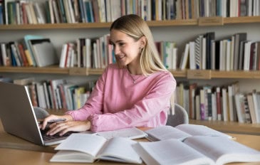 Menina estudando com livros abertos espalhados em uma mesa enquanto digita no notebook