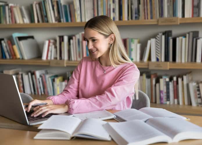 Menina estudando com livros abertos espalhados em uma mesa enquanto digita no notebook